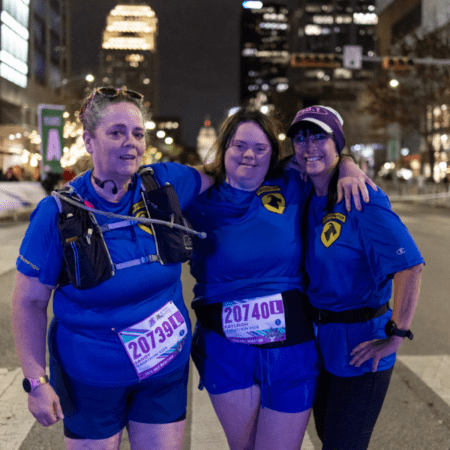 Three women in matching blue shirts and race bibs stand arm-in-arm at a nighttime city race event, representing their company’s Austin Marathon team program, smiling at the camera with tall buildings and bright lights in the background. Austin Marathon Half Marathon & 5K