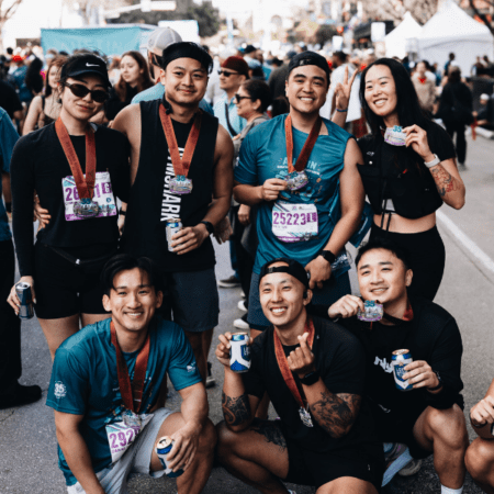 A group of seven smiling runners pose together outdoors after a race, wearing medals and race bibs around their necks. Proud members of the Austin Marathon team program, they celebrate team perks and rewards with drinks near race banners. Austin Marathon Half Marathon & 5K