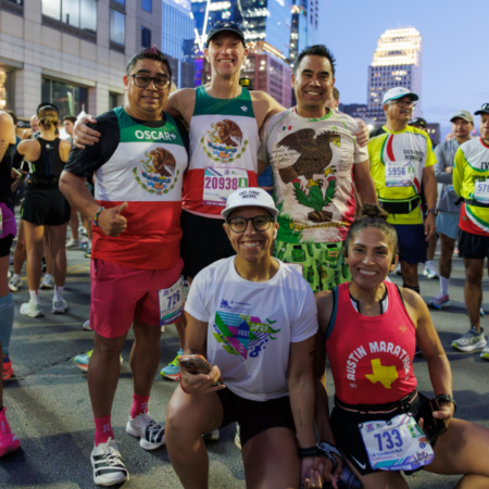 A group of five smiling runners pose together before a race, sporting athletic gear and race bibs. They're participating in the Austin Marathon team program, with a cityscape and fellow Team Registration runners in the background. Austin Marathon Half Marathon & 5K