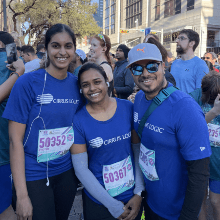 Three people in matching blue "CIRRUS LOGIC" shirts and race bibs smile at the camera in a crowd of runners, showing their excitement as part of the Austin Marathon team program and ready to enjoy team perks and rewards. Austin Marathon Half Marathon & 5K