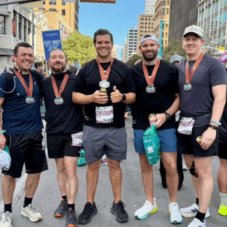 Five men wearing race medals and athletic gear stand together and smile for a photo on a city street after the race, celebrating their Austin Marathon team program success. Buildings and other runners are visible in the background. Austin Marathon Half Marathon & 5K