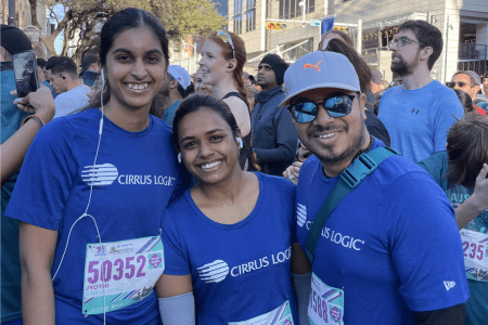 Three people wearing matching blue "Cirrus Logic" shirts smile at the camera during a race event, representing Corporate Teams. Two have race bibs pinned to their shirts, surrounded by other participants in an outdoor city setting. Austin Marathon Half Marathon & 5K