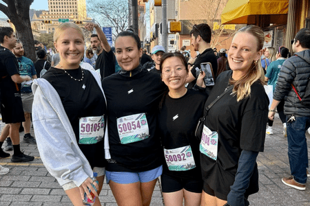 Four women wearing black shirts and race bibs smile and pose together at an outdoor race event—perfect for team building. Other participants and spectators are visible in the background on a bustling city street. Austin Marathon Half Marathon & 5K