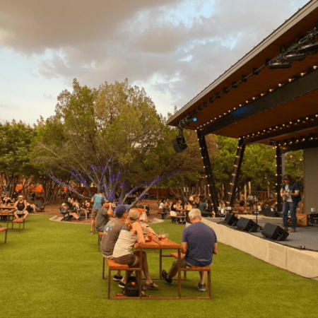 People sit at picnic tables on a grassy lawn, enjoying live music on an outdoor stage under string lights—one of the best patios for a relaxed evening among Austin patio spots, with trees and a cloudy sky in the background. Austin Marathon Half Marathon & 5K