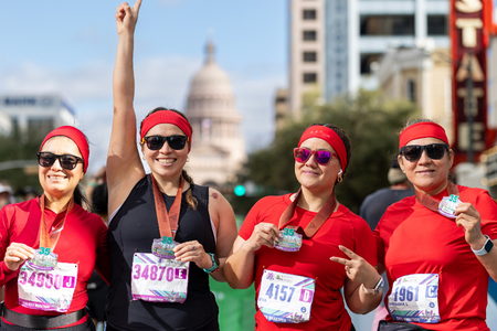 Four women wearing red shirts and headbands proudly display medals after a charity race, smiling for the camera. One woman raises her arm in celebration. City buildings and the Texas State Capitol are visible in the background. Austin Marathon Half Marathon & 5K