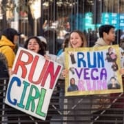 Two women smile and cheer at a destination race in Austin, holding colorful handmade signs that say “RUN GIRL” and “RUN VEGA RUN.” Other spectators stand nearby behind a metal barrier. Austin Marathon Half Marathon & 5K