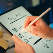 A person uses a stylus to check off items on a digital checklist labeled "PLAN" displayed on a tablet screen, focusing attention on marathon training and running preparation. The blurred background emphasizes the planning activity. Austin Marathon Half Marathon & 5K