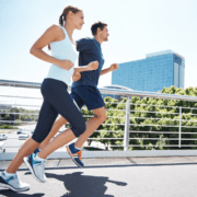 A woman and a man are running outdoors on a sunny day, side by side on a path with city buildings and trees behind them. Dressed in athletic gear, they appear energetic—perhaps in the midst of marathon training. Austin Marathon Half Marathon & 5K