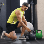 A man in a sleeveless yellow shirt and black shorts kneels on one knee in a gym, reaching for a black kettlebell beside a green one—engaged in base building for marathon training. Exercise equipment and a stability ball are visible in the background. Austin Marathon Half Marathon & 5K