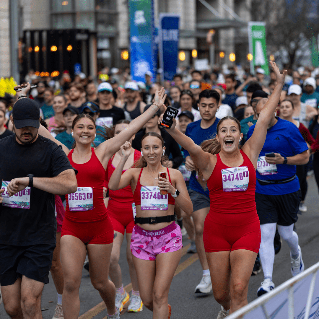 A group of runners, including three women in red outfits, smile and cheer with arms raised as they participate in the Ascension Seton Austin Marathon among a large crowd of competitors on a bustling city street. Austin Marathon Half Marathon & 5K