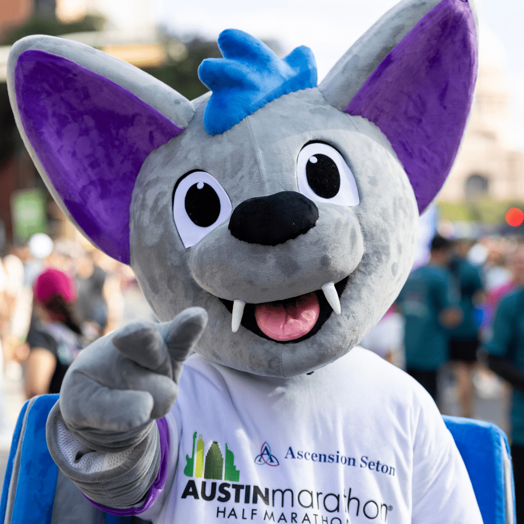A person in a large, gray bat mascot costume with purple ears and blue accents smiles and points at the camera, proudly sporting an "Ascension Seton Austin Marathon Half Marathon" T-shirt at an outdoor event. Austin Marathon Half Marathon & 5K