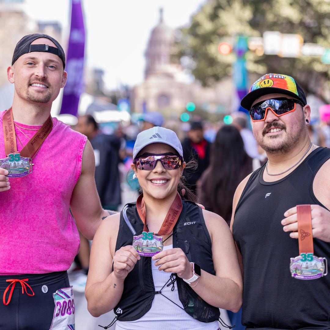 Three runners smile and show off their medals after completing the Ascension Seton Austin Marathon, standing together outdoors with a crowd and city buildings in the background. Austin Marathon Half Marathon & 5K