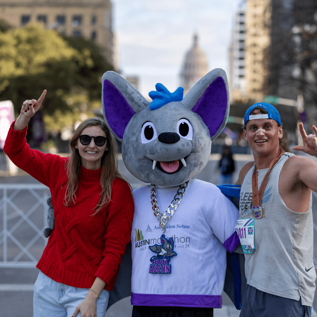 A woman in a red sweater and a man in athletic wear pose with a large bat mascot at an outdoor event, celebrating Austin Marathon history. All three are smiling and making a "number one" gesture against the cityscape and domed building backdrop. Austin Marathon Half Marathon & 5K