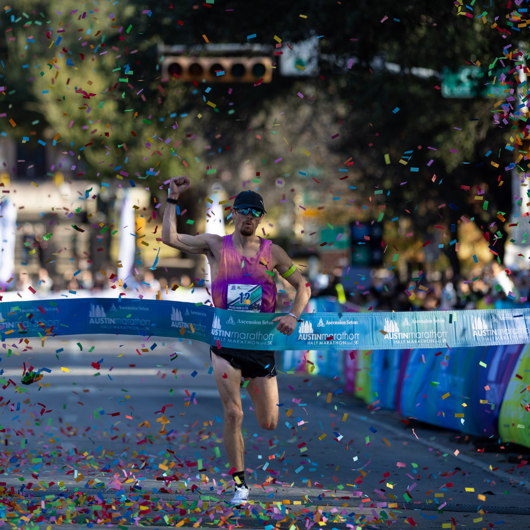 A runner in a pink tank top celebrates crossing the Austin Marathon finish line, surrounded by colorful confetti, with blurred spectators and banners highlighting the event’s rich history in the background. Austin Marathon Half Marathon & 5K
