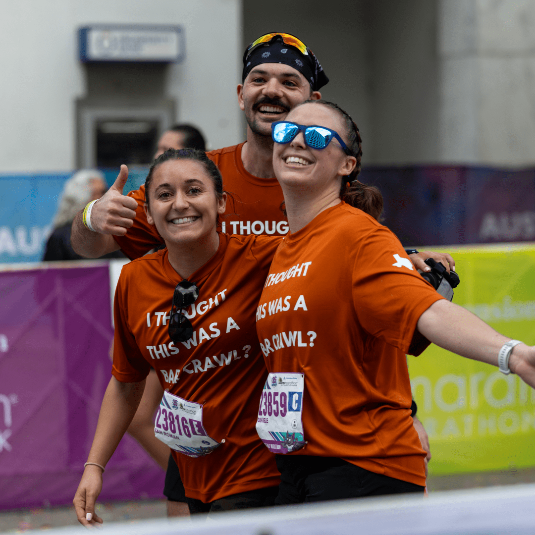 Three smiling runners in matching orange shirts pose at the Ascension Seton Austin Marathon finish line, two women in front and a man behind giving a thumbs-up. Their shirts humorously read, “I thought this was a bar crawl?!”. Austin Marathon Half Marathon & 5K