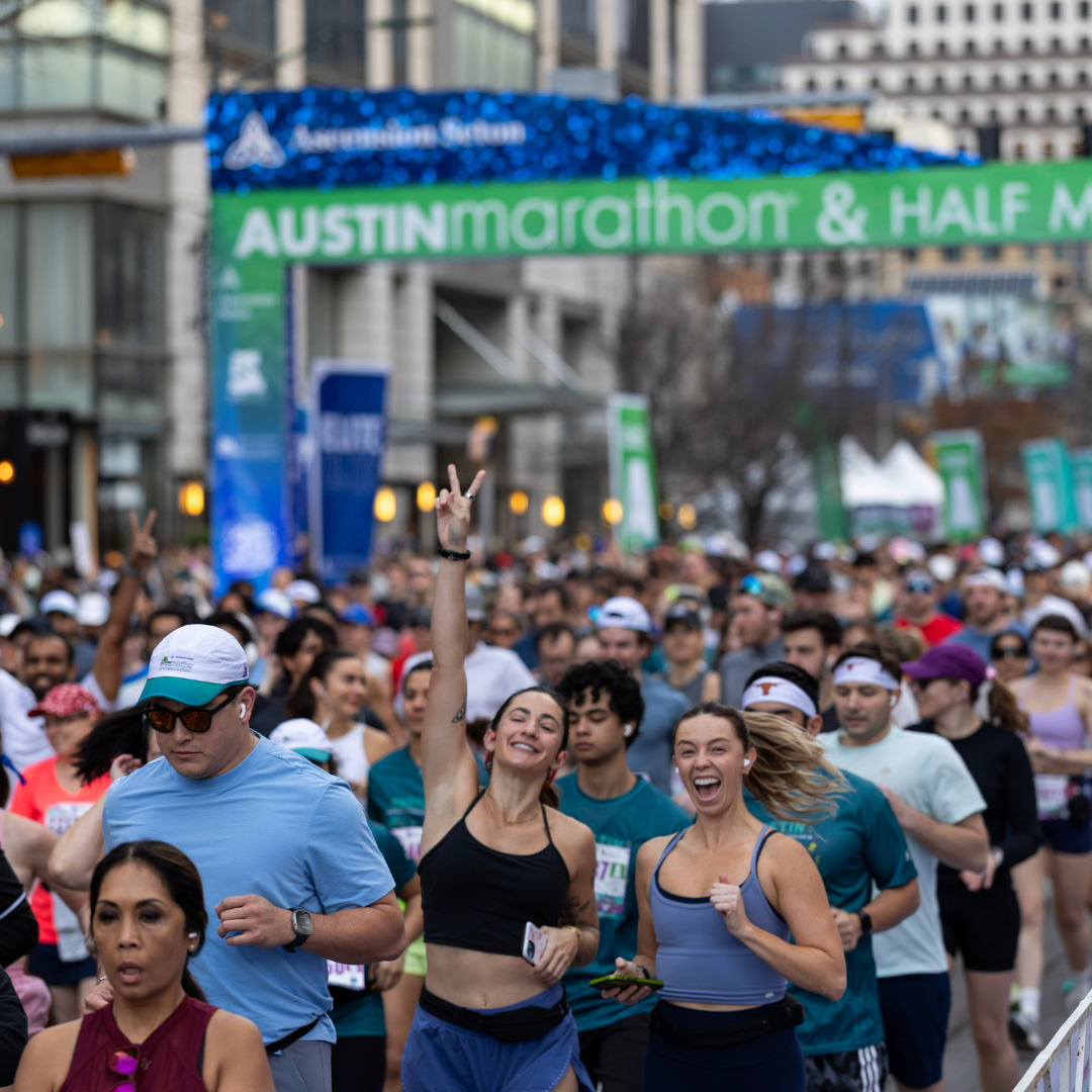 A large group of runners participates in the Ascension Seton Austin Marathon & Half Marathon. Two women at the front smile and gesture enthusiastically, while others follow behind under the event’s green and blue banner. Austin Marathon Half Marathon & 5K