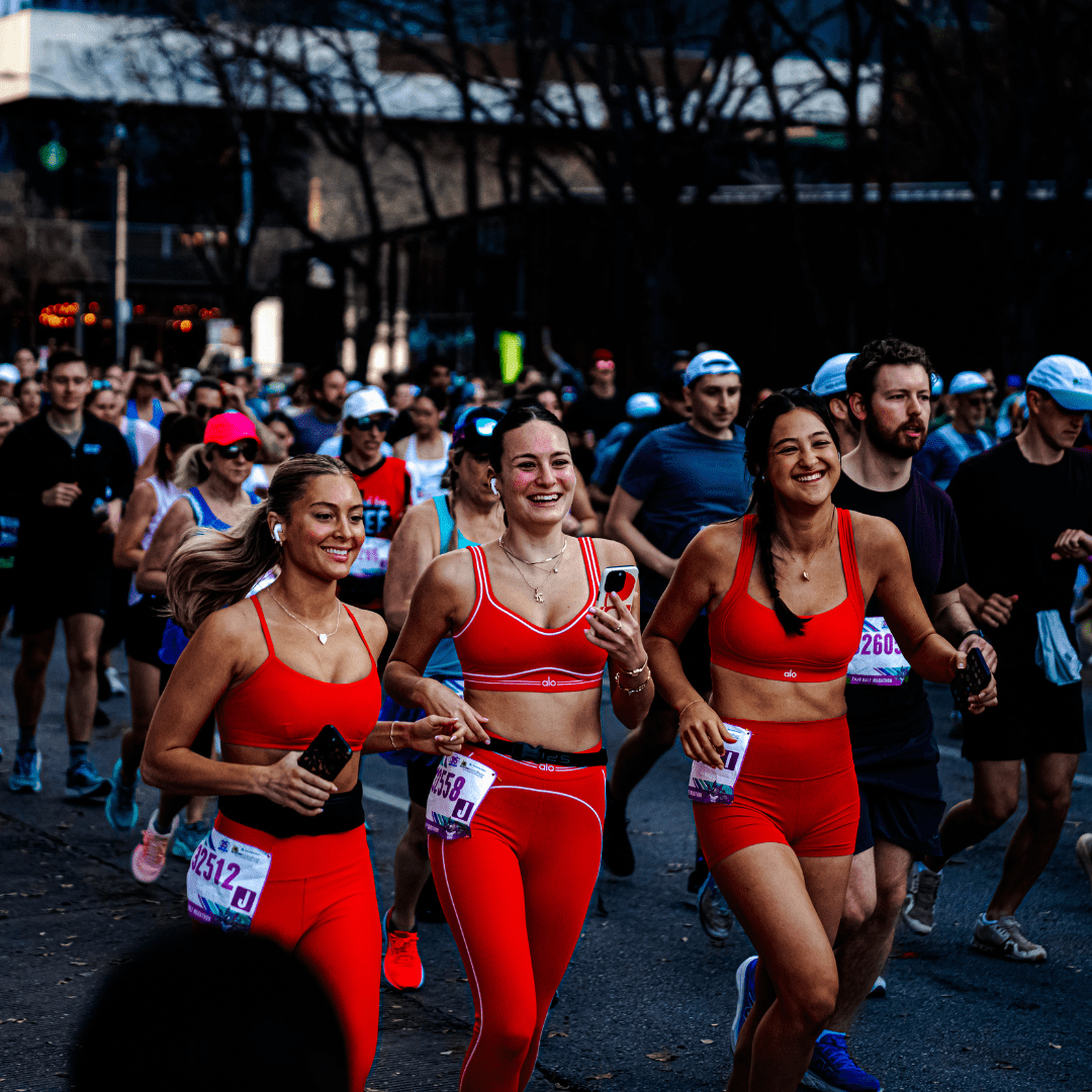 Three women in matching red athletic outfits run and smile together during the Ascension Seton Austin Marathon, surrounded by other racers. All wear bibs and seem to be enjoying the event, with trees and city buildings in the background. Austin Marathon Half Marathon & 5K