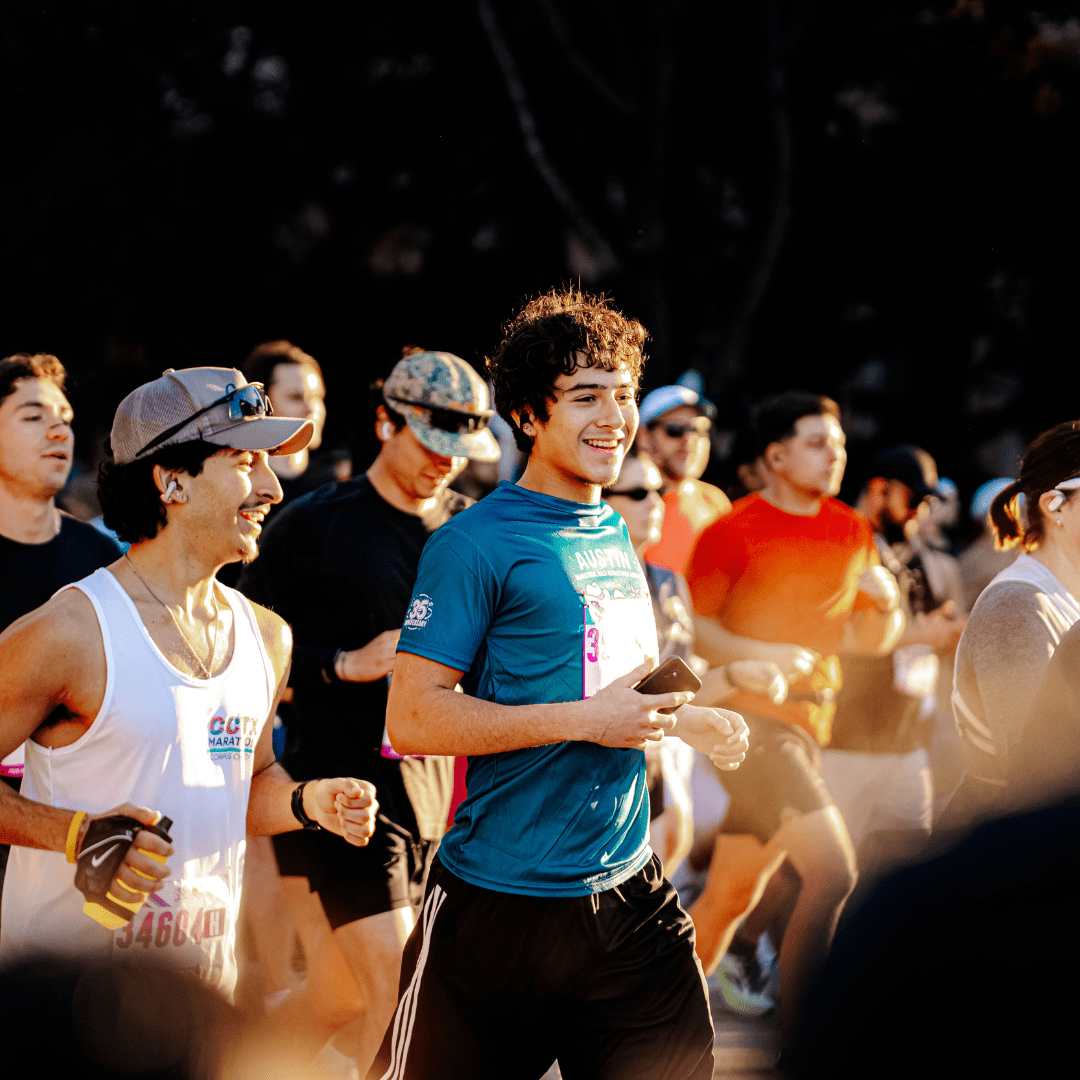 A group of people run outdoors in the Austin Marathon, with two young men in the foreground smiling and wearing athletic gear and race bibs, surrounded by other runners in sunlight, capturing the excitement of Austin Marathon history. Austin Marathon Half Marathon & 5K