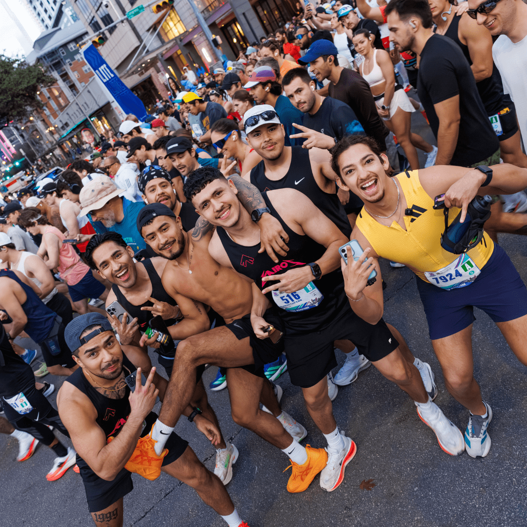 A group of smiling runners pose for a photo at the crowded Ascension Seton Austin Marathon starting line, wearing athletic gear and race bibs, with excited participants and tall buildings in the background. Austin Marathon Half Marathon & 5K