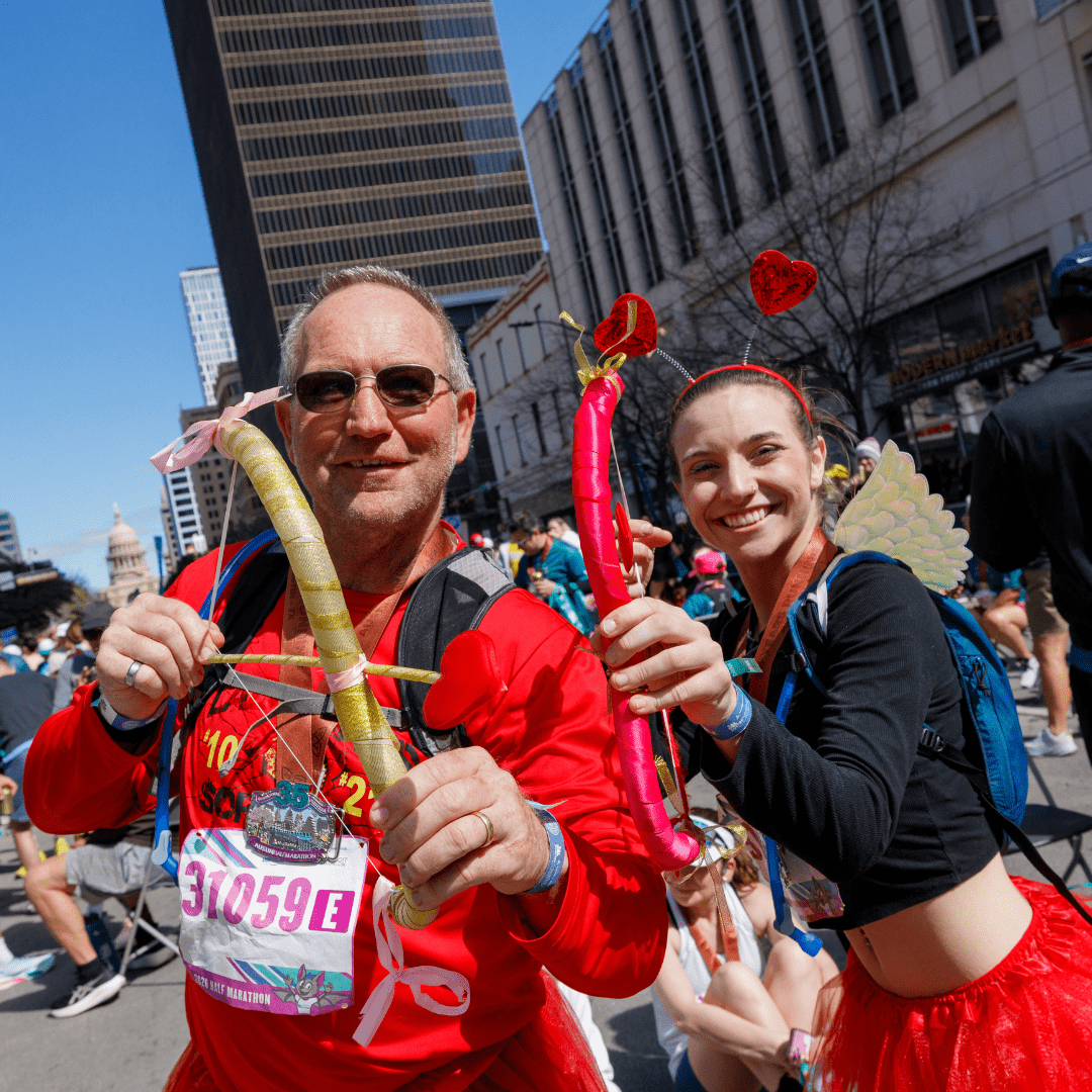 Two people in red costumes with heart-themed accessories and balloon swords smile at a street event, with crowds, tall buildings, and the Texas State Capitol in the background—capturing the festive spirit of Austin Marathon registration day. Austin Marathon Half Marathon & 5K