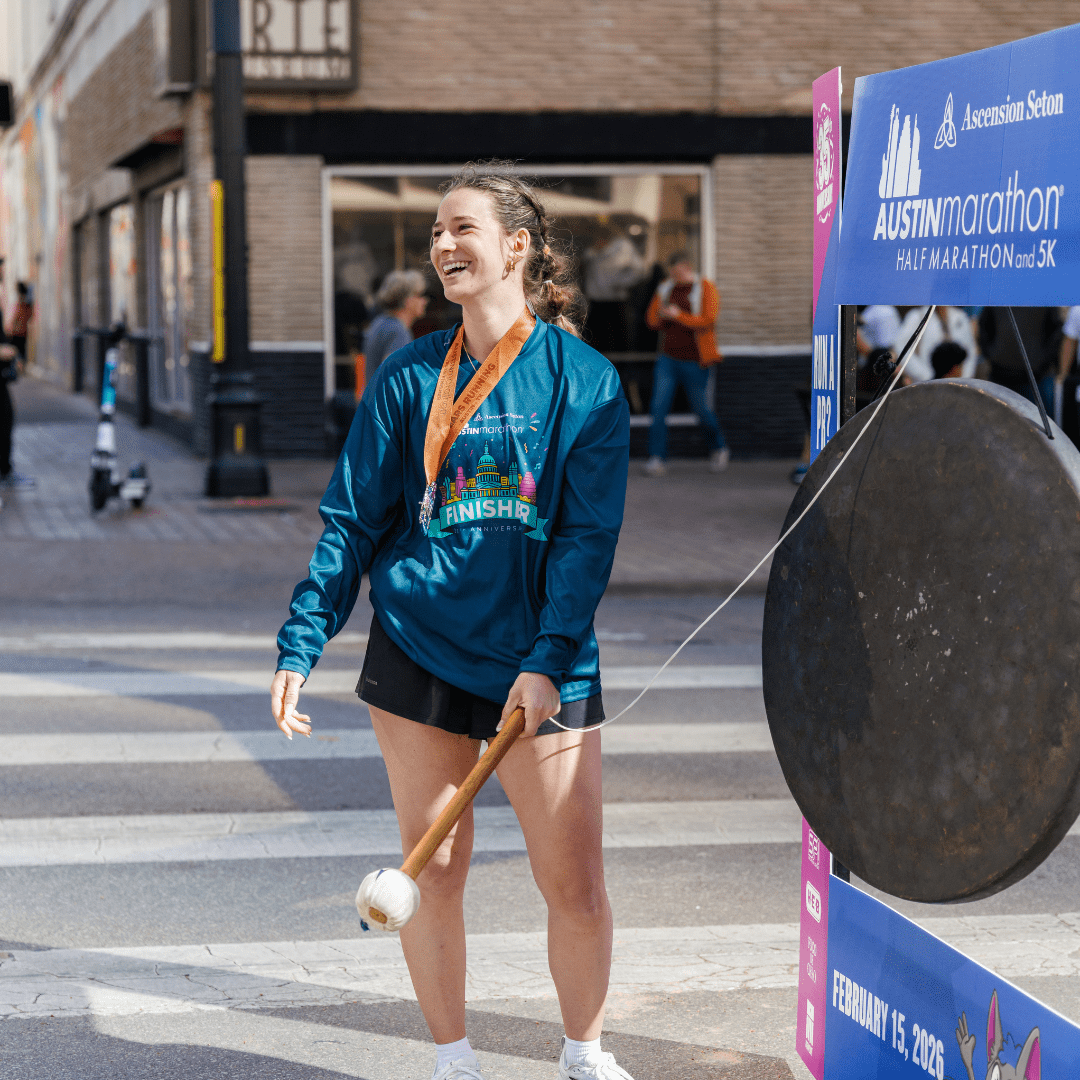 A smiling woman wearing a race finisher medal and shirt stands at the Ascension Seton Austin Marathon finish line, holding a mallet and about to strike a large gong. She appears happy and triumphant. Austin Marathon Half Marathon & 5K