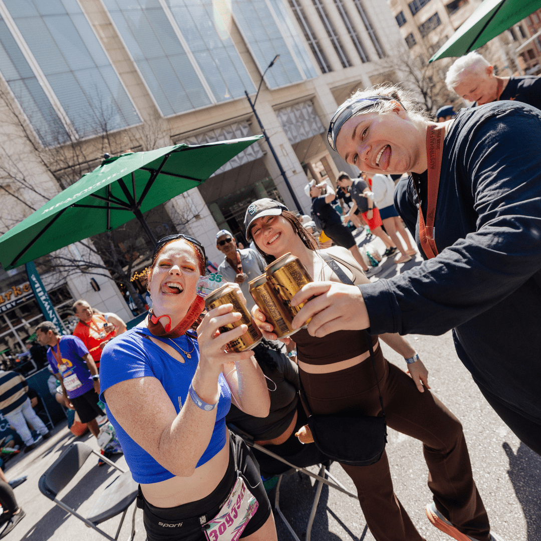 Three smiling women in athletic wear hold up drinks and pose for a photo at the Ascension Seton Austin Marathon, with people and umbrellas in the background on a sunny day in the city. Austin Marathon Half Marathon & 5K
