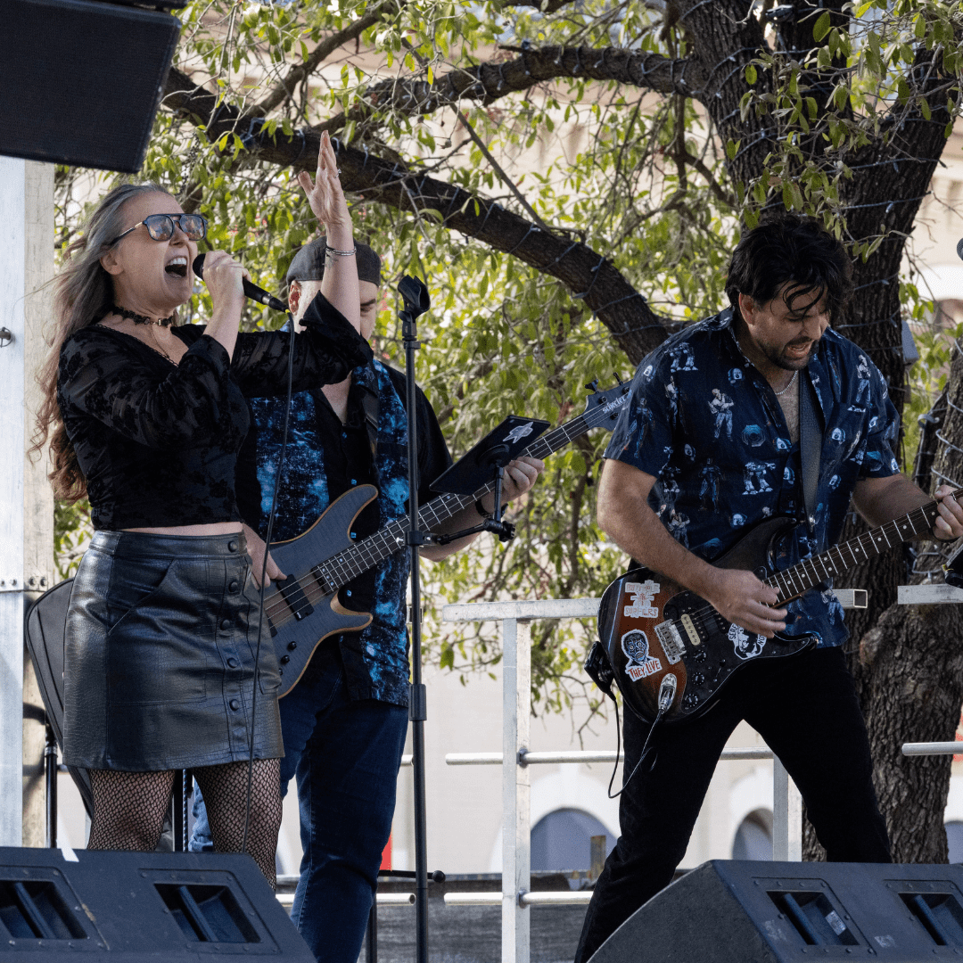 A woman passionately sings into a microphone while two men play electric guitar and bass on an outdoor stage, as leafy trees and lights set the scene—capturing the vibrant energy that echoes throughout Austin Marathon history. Austin Marathon Half Marathon & 5K