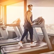 A person wearing headphones, a sleeveless shirt, and athletic pants walks on a treadmill in a gym with large windows, as sunlight streams in—perfect for picking up summer running tips. Austin Marathon Half Marathon & 5K