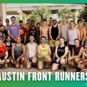 A group of about 20 smiling runners from the Buda Running Club pose together outdoors under a bridge, some standing and some kneeling. They wear athletic gear, with “AUSTIN FRONT RUNNERS” text on a green background. Austin Marathon Half Marathon & 5K
