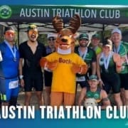A group of triathletes, some wearing medals, pose and smile in front of an "Austin Triathlon Club" tent with a person in a moose mascot costume named Buck at the center during a fun Group Run event. Austin Marathon Half Marathon & 5K