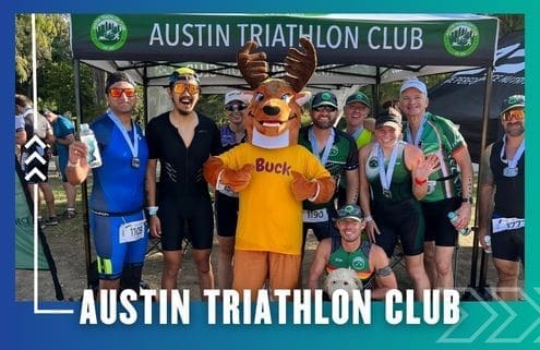 A group of triathletes, some wearing medals, pose and smile in front of an "Austin Triathlon Club" tent with a person in a moose mascot costume named Buck at the center during a fun Group Run event. Austin Marathon Half Marathon & 5K