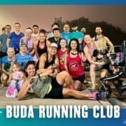 A diverse group of people, including men, women, children, and a stroller, smile and pose together outside at night for a group run under the text "BUDA RUNNING CLUB" with decorative blue and white graphics. Austin Marathon Half Marathon & 5K