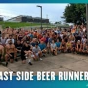 A large group of people in athletic wear pose together outdoors on a paved area, representing the Buda Running Club. "East Side Beer Runners" is written in bold white text at the bottom, with trees and a building visible in the background. Austin Marathon Half Marathon & 5K