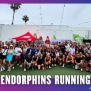 A large group of people in athletic wear pose together outdoors on a sports court under string lights, with a red New Balance tent and surfboards in the background. Text at the bottom reads "ENDORPHINS RUNNING GROUP RUN. Austin Marathon Half Marathon & 5K