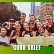 A group of runners from Buda Running Club pose together and smile after a race, displaying their medals. They stand outdoors in a city, holding a "Good Grief" banner with buildings and the finish line in the background. Austin Marathon Half Marathon & 5K