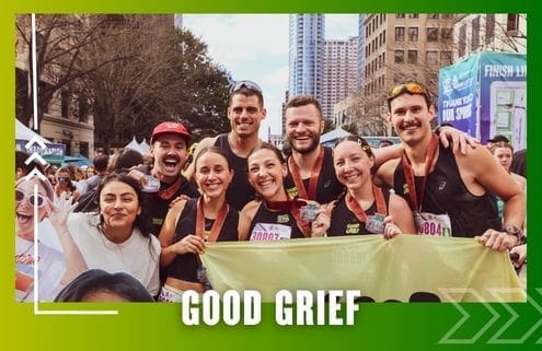 A group of runners from Buda Running Club pose together and smile after a race, displaying their medals. They stand outdoors in a city, holding a "Good Grief" banner with buildings and the finish line in the background. Austin Marathon Half Marathon & 5K