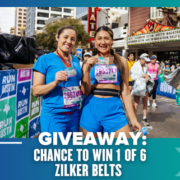 Two smiling women in athletic wear stand holding medals and water bottles at a marathon finish line. Behind them, participants walk by and a banner advertises a Giveaway to win 1 of 6 Zilker Belts. Austin Marathon Half Marathon & 5K