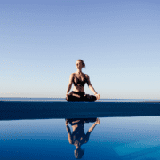 A woman sits cross-legged in a yoga pose by a pool, her reflection visible in the water. Dressed in athletic wear under a clear blue sky, she radiates calm and patience—a moment of peace during her marathon training. Austin Marathon Half Marathon & 5K