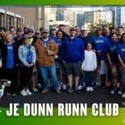 A group of people, some in blue "Run Club" shirts, pose and smile together outdoors in front of buildings. A dog sits in front. The image has a green border and reads "JE DUNN RUNN CLUB," celebrating their latest Group Run. Austin Marathon Half Marathon & 5K
