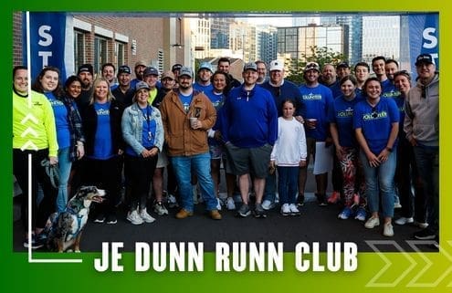 A group of people, some in blue "Run Club" shirts, pose and smile together outdoors in front of buildings. A dog sits in front. The image has a green border and reads "JE DUNN RUNN CLUB," celebrating their latest Group Run. Austin Marathon Half Marathon & 5K