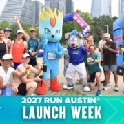 A group of smiling runners pose with two cheerful mascots at the 2027 Run Austin Launch Week event, with city skyscrapers and event banners visible in the background. Austin Marathon Half Marathon & 5K
