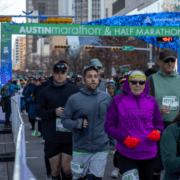 Runners in athletic gear participate in the Austin Marathon & Half Marathon, passing under a blue and green start banner. A woman in a bright pink "Just Do It" shirt leads the way, showcasing the underrated skill of patience gained through marathon training. Austin Marathon Half Marathon & 5K