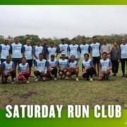 A group of people in matching "Saturday Run Club" shirts pose outdoors on grass, smiling for a group photo after a group run. The image has a green gradient border and text that reads "Saturday Run Club" at the bottom. Austin Marathon Half Marathon & 5K