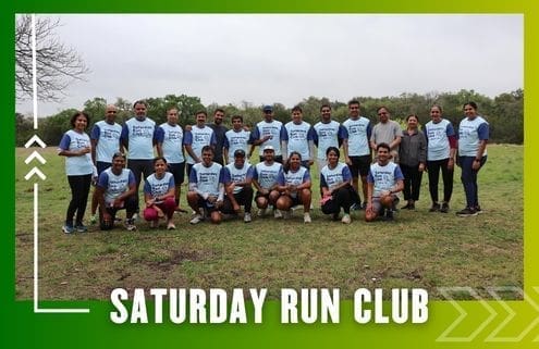 A group of people in matching "Saturday Run Club" shirts pose outdoors on grass, smiling for a group photo after a group run. The image has a green gradient border and text that reads "Saturday Run Club" at the bottom. Austin Marathon Half Marathon & 5K