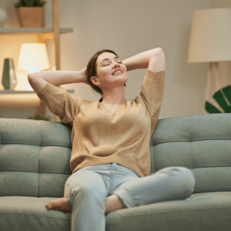 Woman sitting on a green sofa with her hands behind her head, eyes closed and smiling, appearing relaxed—enjoying a moment of calm after overcoming post-race blues. Warm indoor lighting and modern decor are visible in the background. Austin Marathon Half Marathon & 5K