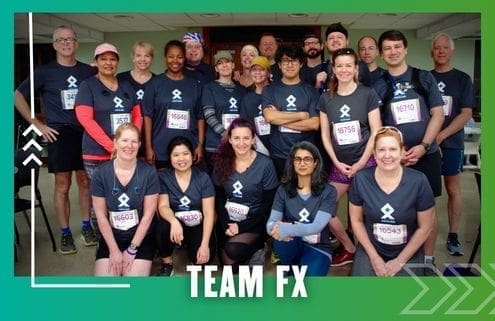 A group of people wearing matching "TEAM FX" shirts and race bibs pose together indoors, smiling, in front of a green and blue graphic border with "TEAM FX" text at the bottom, celebrating their Group Run. Austin Marathon Half Marathon & 5K
