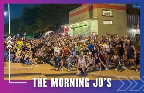 A large, diverse group of people—possibly from a Group Run or the Buda Running Club—poses together outside a building at night under the sign "I love you." Text at the bottom reads "THE MORNING JO’S" on a purple and blue border. Austin Marathon Half Marathon & 5K