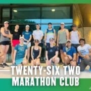 A group of fourteen people in athletic attire pose together outside a building, representing the Buda Running Club. Text at the bottom reads "TWENTY-SIX TWO MARATHON CLUB" on a green border with arrow graphics. Austin Marathon Half Marathon & 5K