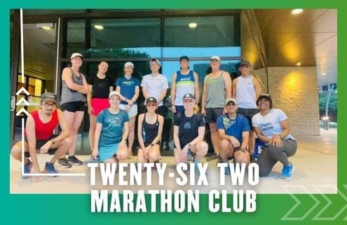 A group of fourteen people in athletic attire pose together outside a building, representing the Buda Running Club. Text at the bottom reads "TWENTY-SIX TWO MARATHON CLUB" on a green border with arrow graphics. Austin Marathon Half Marathon & 5K