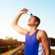 A young man in a blue tank top pours water over his head from a plastic bottle while standing outdoors on a sunny day, embodying summer fitness and appearing hot and sweaty from physical activity. Austin Marathon Half Marathon & 5K